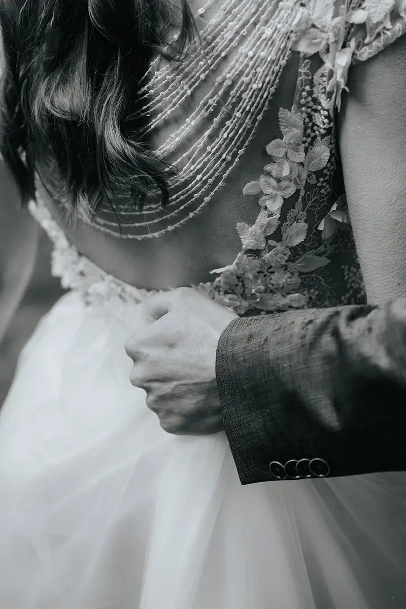 hochzeit-hotel-bedburger-muehle-hochzeitsfotograf-philipp-wildfeuer-brautkleid-halten Hochzeit Hotel Bedburger Mühle – Schwarzweißaufnahme, in der der Bräutigam das Kleid der Braut mit Spitzenrücken festhält, aufgenommen von Hochzeitsfotograf Philipp Wildfeuer.