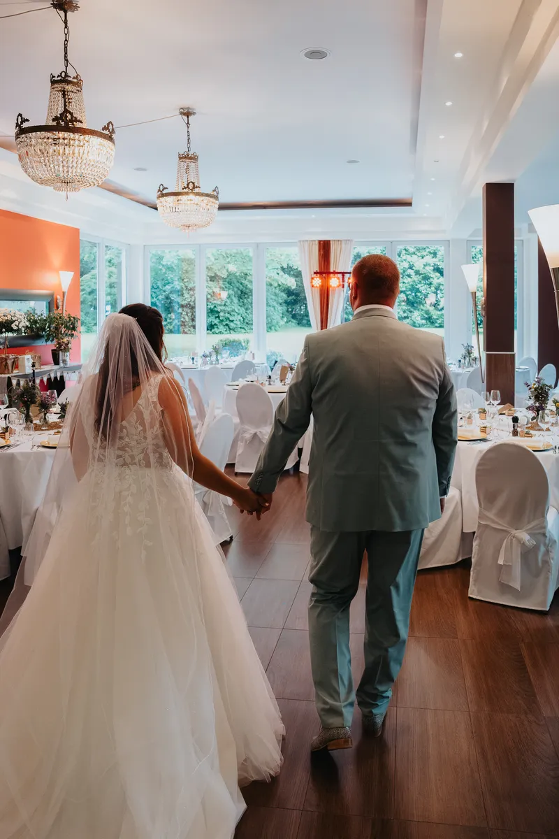 Das Brautpaar betritt Hand in Hand den festlich geschmückten Saal unter funkelnden Kronleuchtern; aufgenommen vom Hochzeitsfotograf Neunkirchen‑Seelscheid.