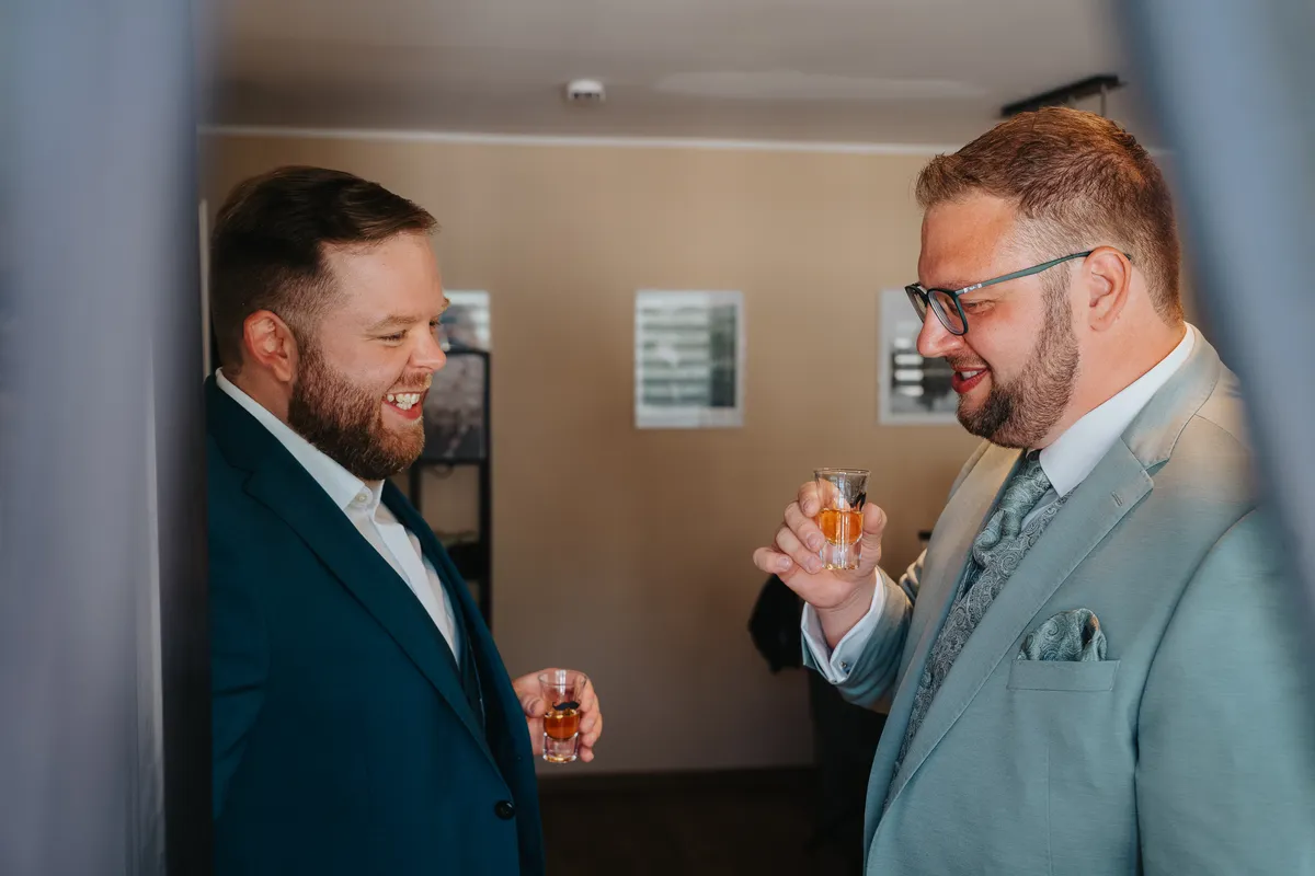 Hochzeitsfotograf Neunkirchen-Seelscheid: Bräutigam und Trauzeuge stehen sich gegenüber, lachen und haben ein Schnapsglas in der Hand.